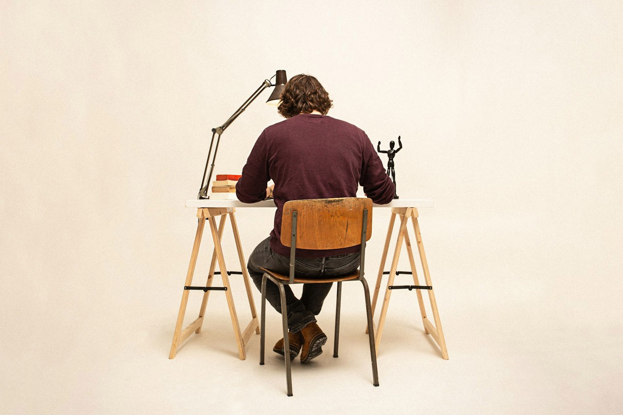 A man sitting and writing at a small wooden desk in a beige room, photo taken from behind.