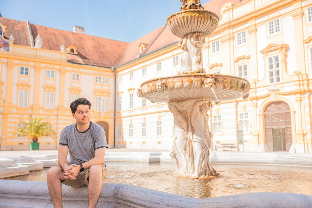 A photo of Jay, a short-haired man, sitting by an ornate white fountain, wearing a grey t-shirt and brown shorts, hands clasped, looking up.